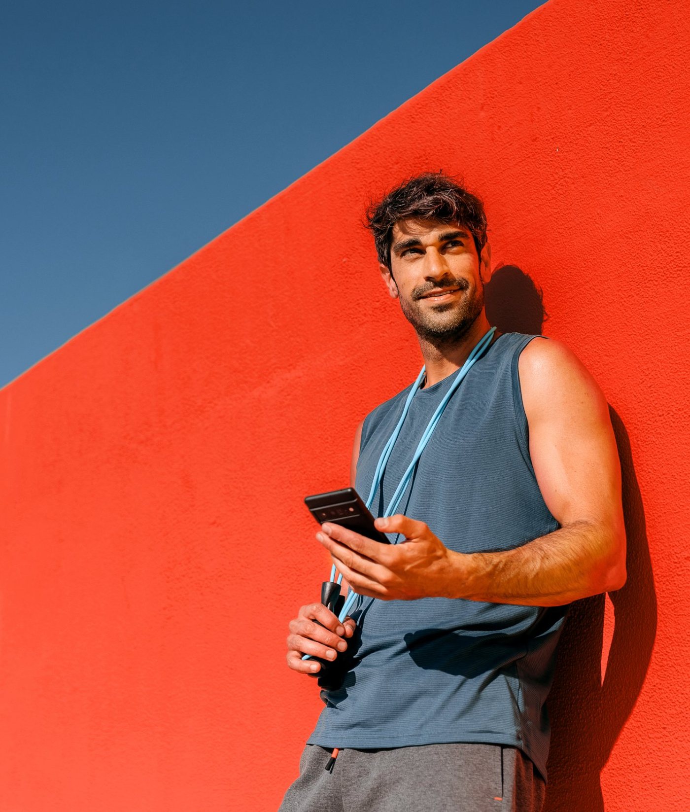 A man leaning on a red wall in the sun, phone in hand